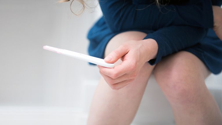 A woman is waiting for the result of a pregnancy test, which she is holding in her hands. She is sitting on the toilet in a bathroom.