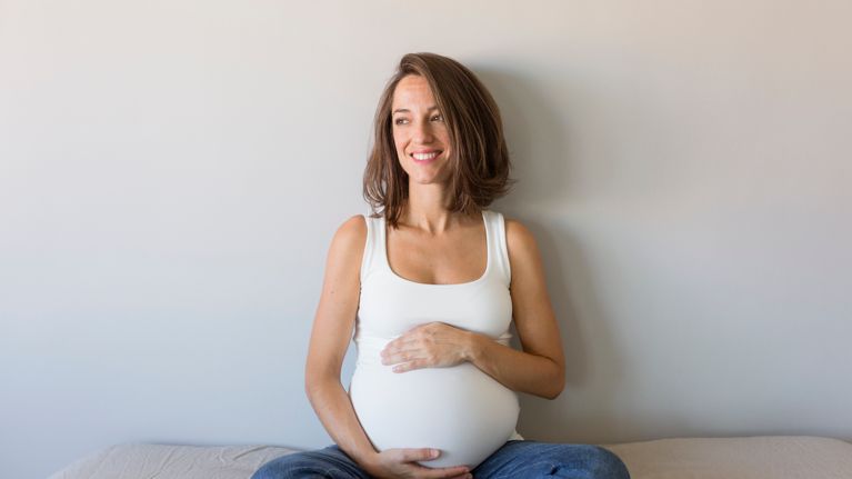 Smiling Pregnant Woman Touching Belly While Sitting On Bed At Home