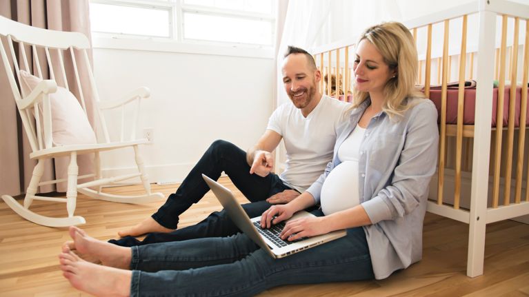 A happy man and his pregnant wife with computer on the baby room at home