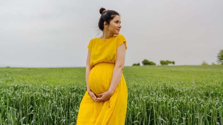 Photo of a pregnant woman standing alone on a grass field, stroking her baby bump and enjoying the calm meadow.