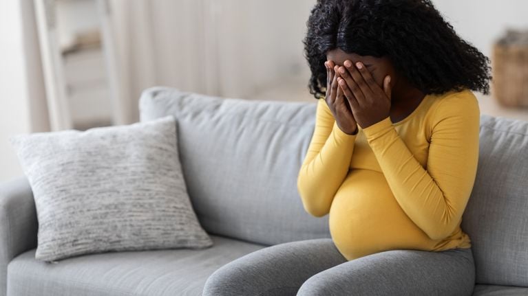 ung pregnant woman feeling sad and crying at home, sitting on couch at living room alone, 