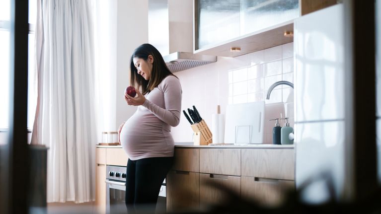 Smiling young pregnant woman touching her belly, eating an fresh organic apple in kitchen at home.