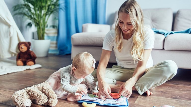 A babysitter with a 1-year-old girl is reading a children's playbook at home on the floor.