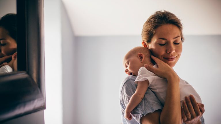 A happy new mother smiles as she craddles her sleeping baby in her arms. She has her eyes closed as she holds her close.