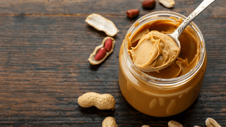 jar of peanut butter sitting on a table with a spoon and loose peanuts next to it