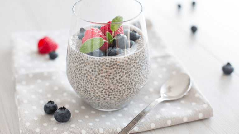 cup of chia pudding with blueberries and strawberries on top and loose berries and a spoon next to it on a table