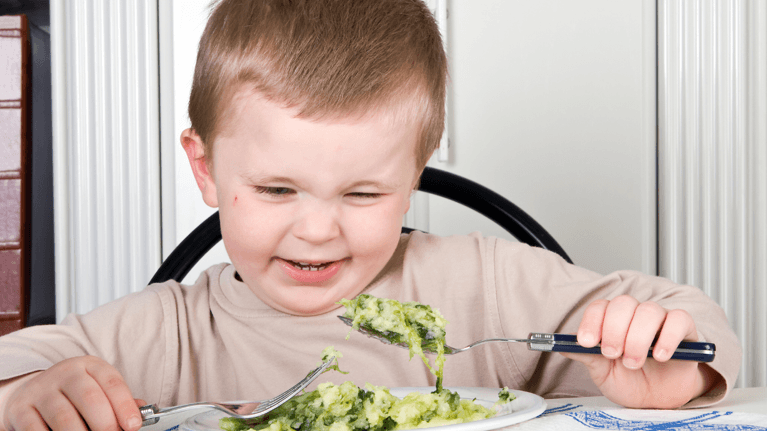 young boy sitting at a plate of vegetables holding up a fork and frowning