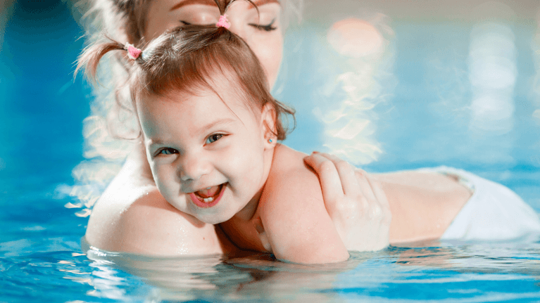mom holding baby in a pool while she looks at the camera smiling
