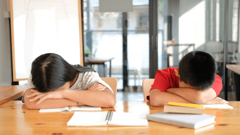 two kids sitting at a kitchen table, heads down on books as if they have fallen asleep