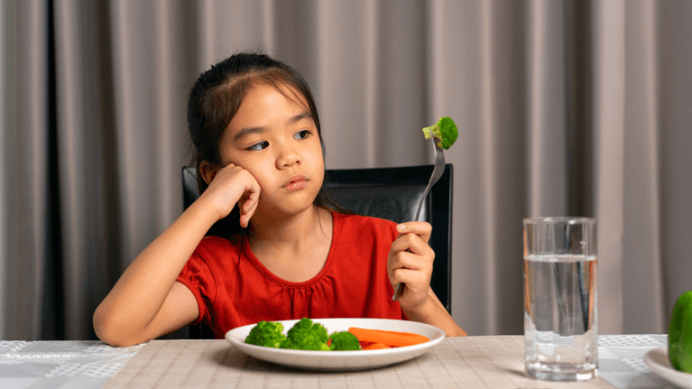 young girl sitting in front of a plate of vegetables frowning