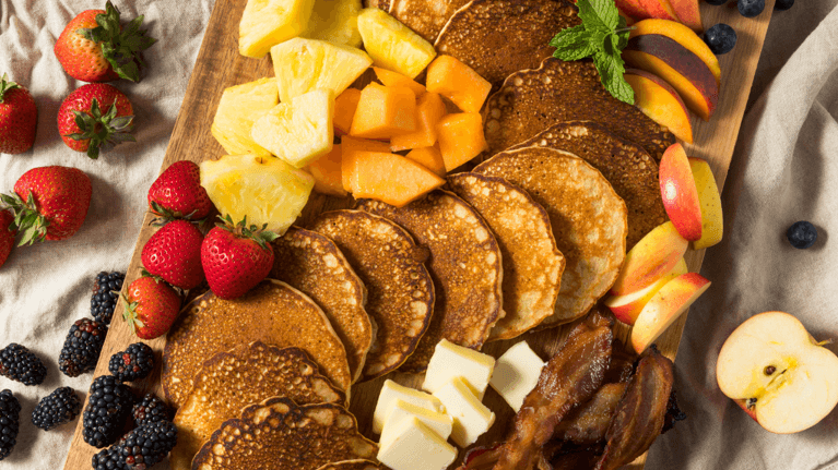 close up photo of a charcuterie board with pancakes, bacon and different fruit on it