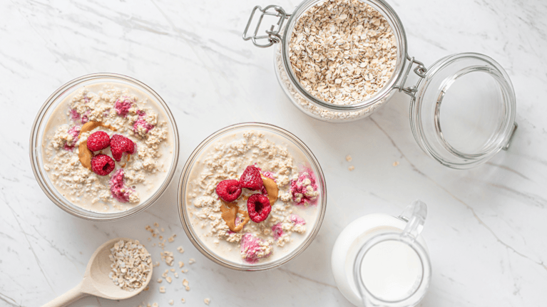 two jars of overnight oats with a jar of oats, wooden spoon and small jug of milk next to it