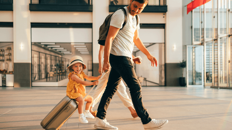 Dad walking through an airport pulling a suitcase while his daughter rides on it, both smiling